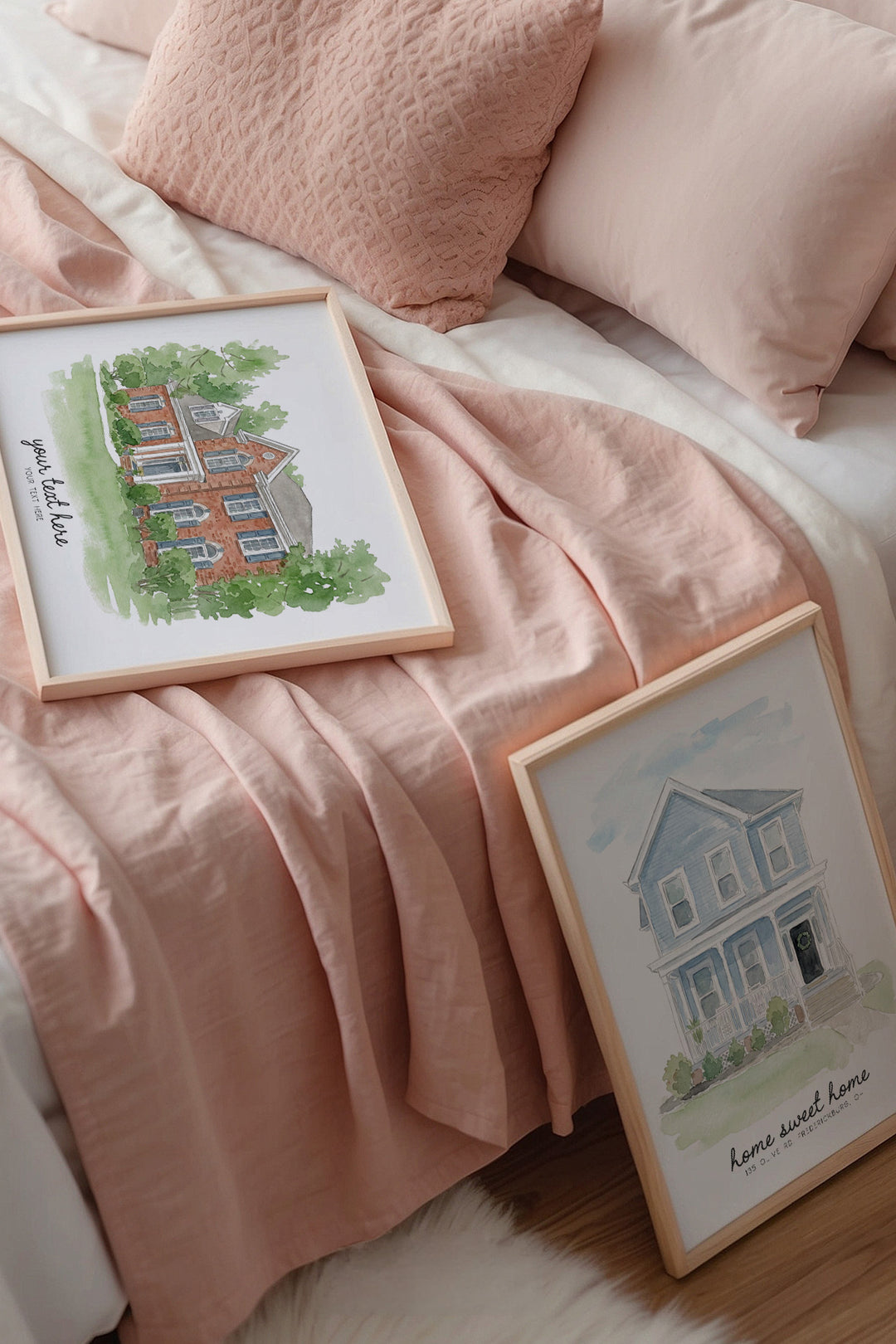 Two framed watercolor house art prints styled on a bed with pink bedding and pillows. One print shows a red brick house and the other shows a blue house, both in light wood frames.
