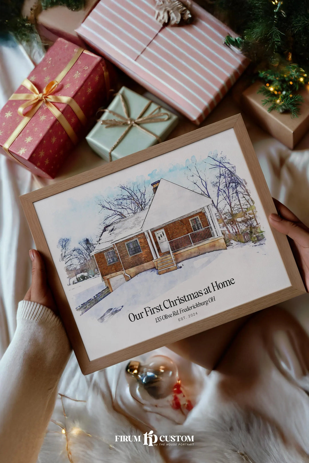 A framed custom watercolor Christmas print held by hands, surrounded by wrapped Christmas gifts and holiday decorations. The print features a watercolor illustration of a cozy brick house on a snowy winter landscape with the text 'Our First Christmas at Home EST.

