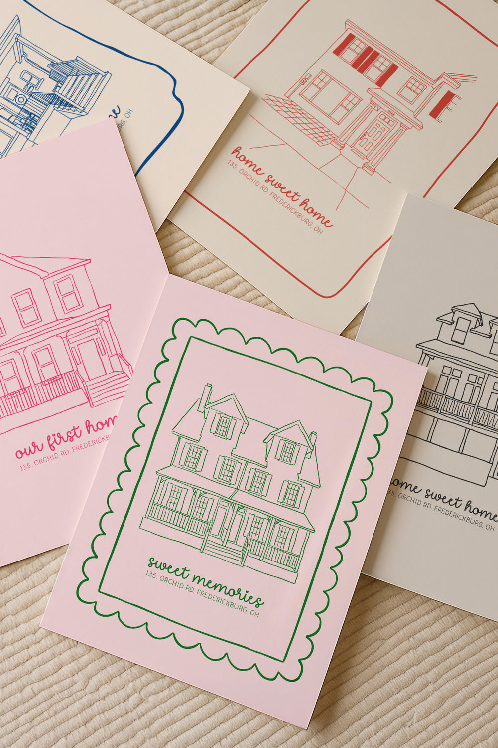 A set of illustrated house-themed cards or prints arranged on a textured cream fabric background. The visible designs include pink, red, blue, and black line drawings of houses with phrases such as "our first home," "home sweet home," and "sweet memories.