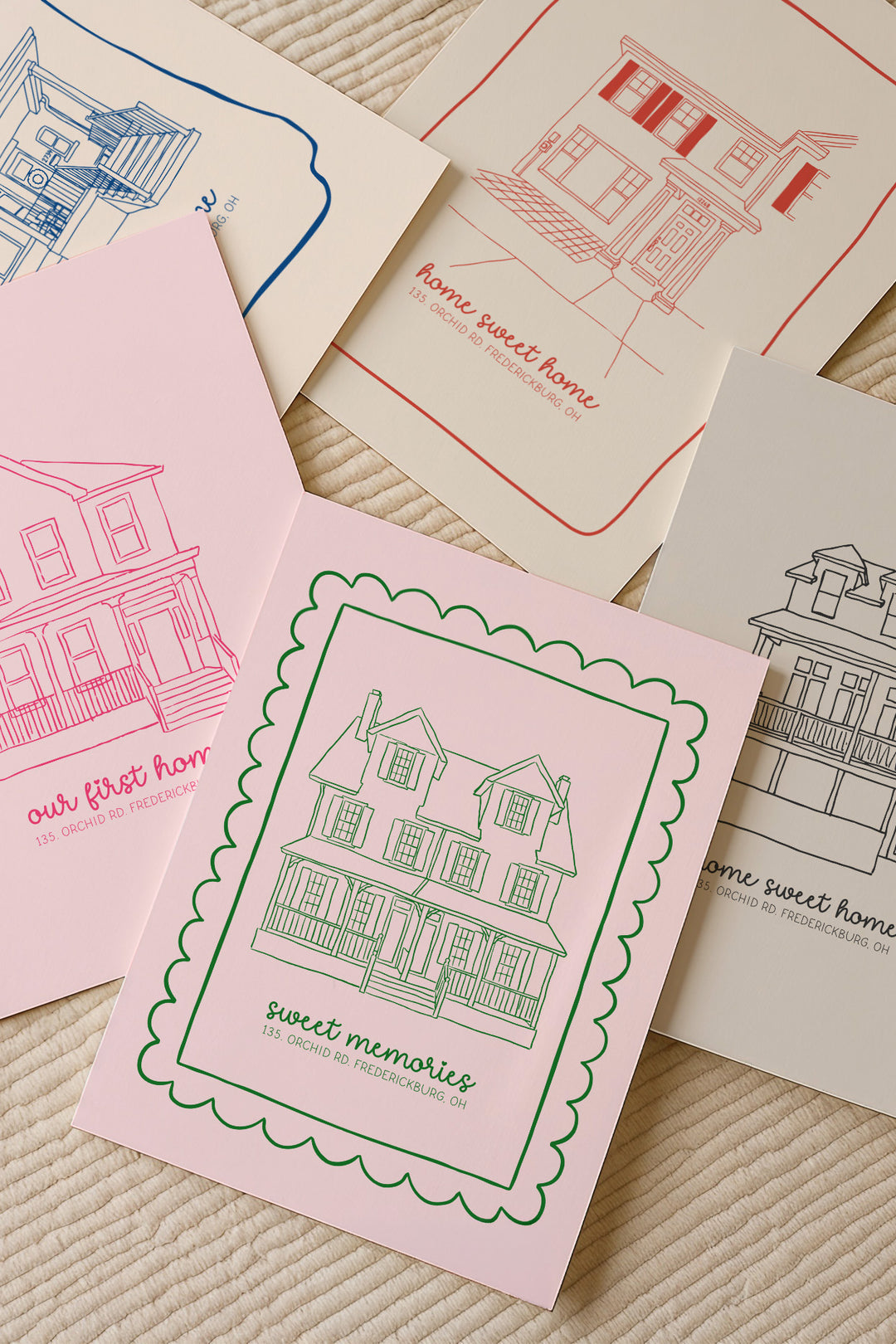 A set of illustrated house-themed cards or prints arranged on a textured cream fabric background. The visible designs include pink, red, blue, and black line drawings of houses with phrases such as "our first home," "home sweet home," and "sweet memories.