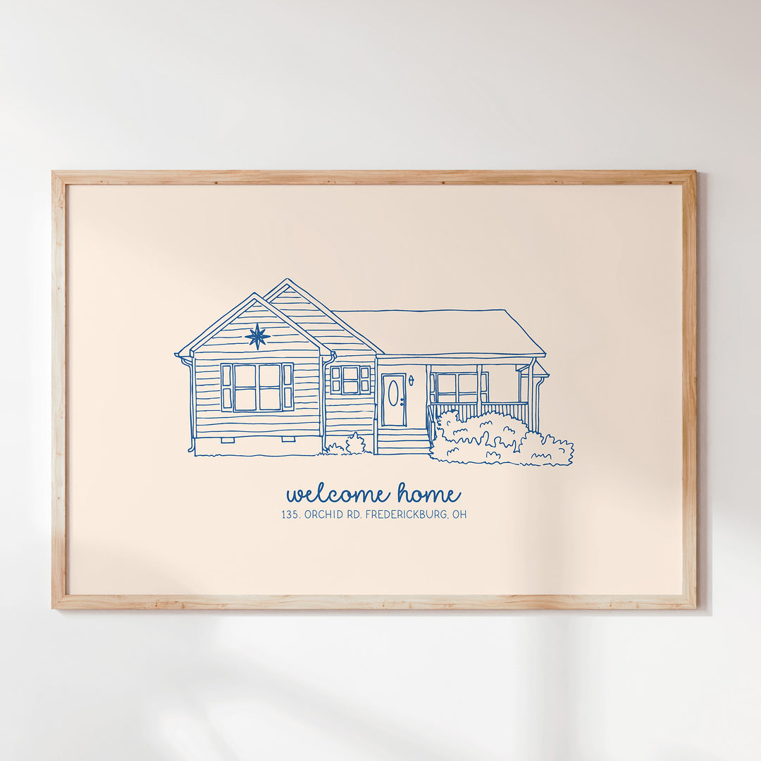 A framed art print featuring a blue line drawing of a house with the text 'welcome home' and '135 Orchard Rd Fredericksburg, OH' on a cream background, enclosed in a light wood frame and mounted on a white wall.
