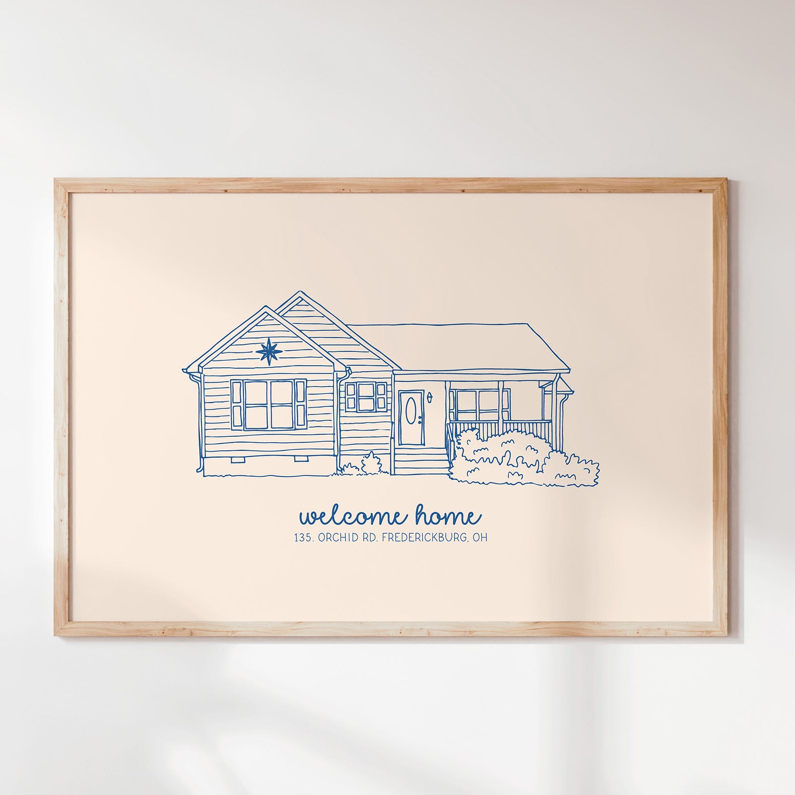 A framed art print featuring a blue line drawing of a house with the text 'welcome home' and '135 Orchard Rd Fredericksburg, OH' on a cream background, enclosed in a light wood frame and mounted on a white wall.