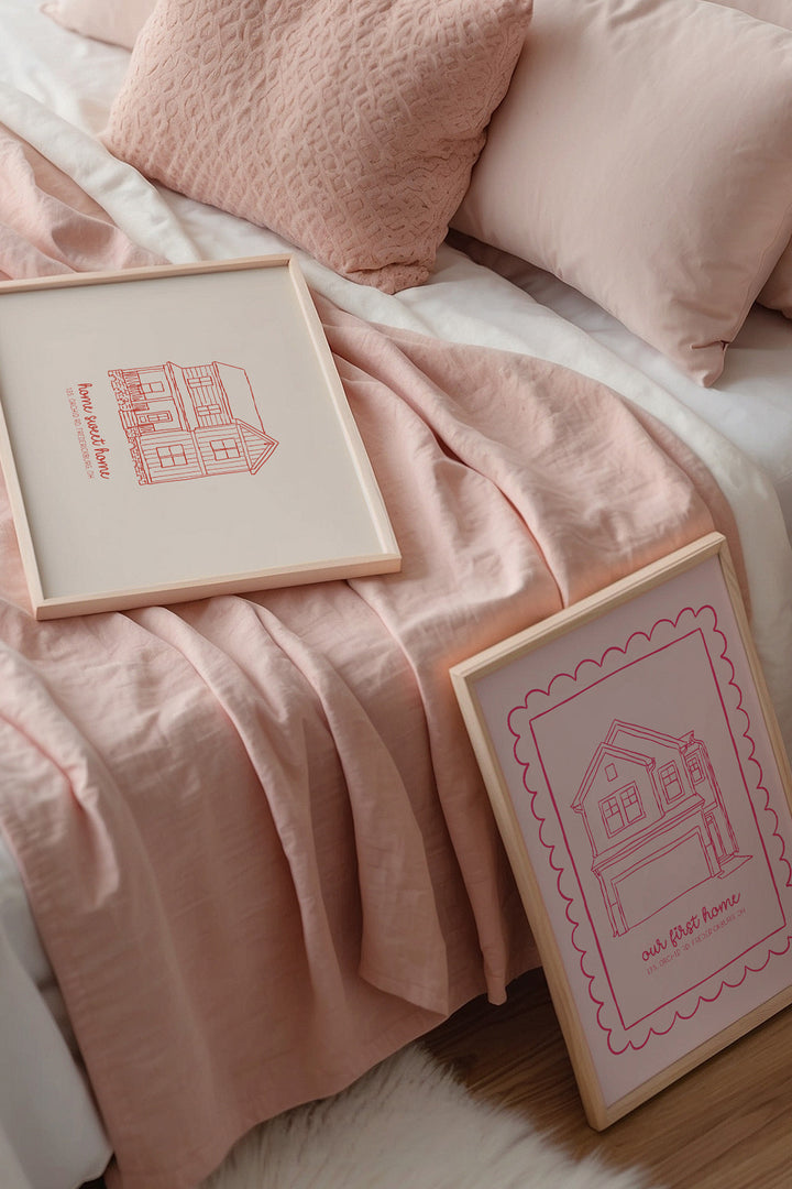 Two framed art prints with pink house illustrations are styled on a bed with blush pink bedding and pillows.