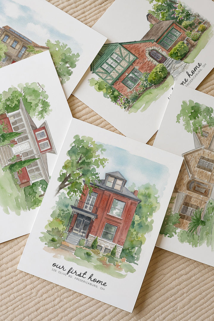 A set of watercolor art prints depicting different red brick houses, arranged in a flat-lay pattern on a beige textured blanket. The central print reads 'our first home' with a small address below.