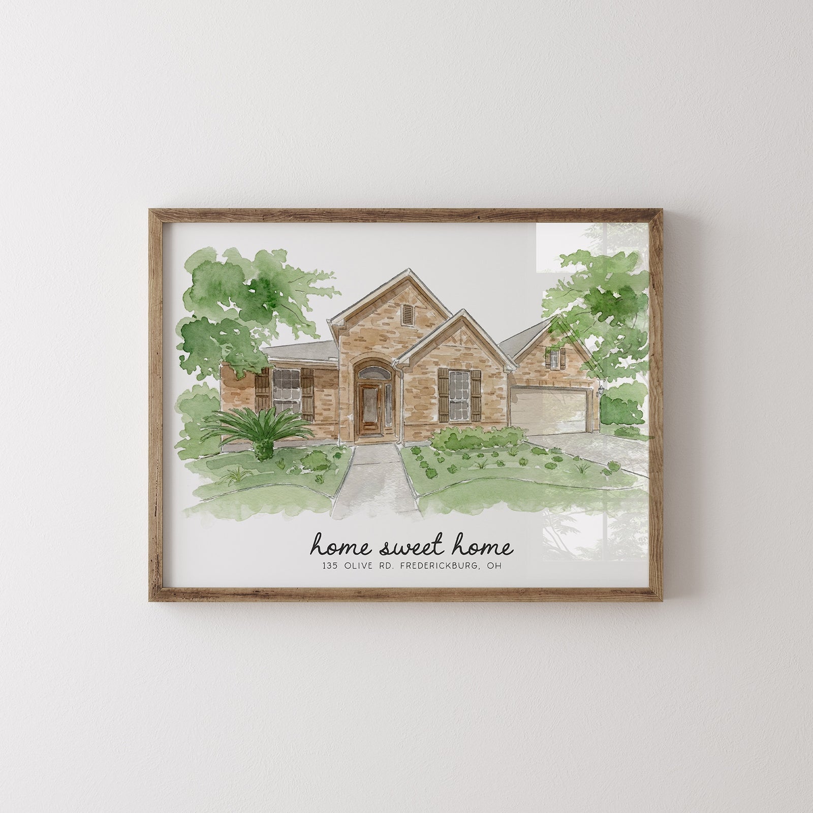 A framed watercolor art print of a suburban house with the text 'home sweet home' and a small address, shown hanging on a light gray wall.